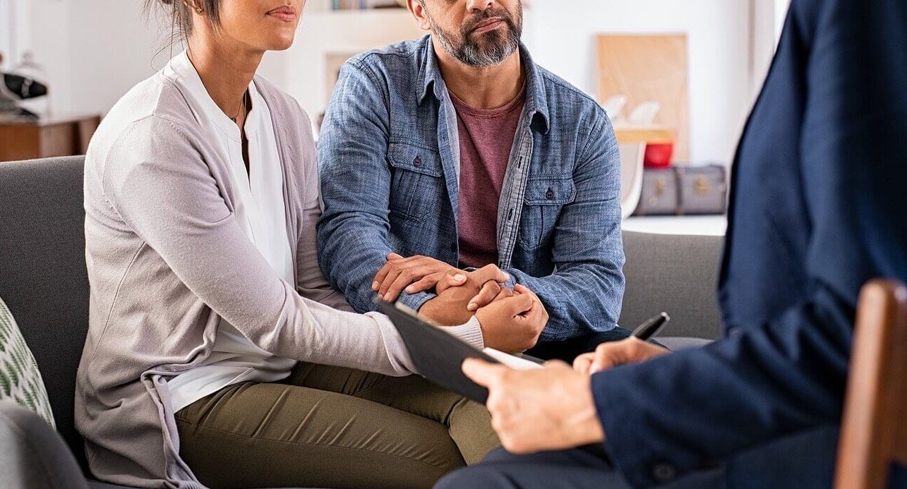 Image of a couple holding hands while sitting on a couch listening to a therapist. With the support of EMDR therapy in Wake Forest, NC you and your partner can begin to rebuild trust after betrayal and start healing your relationship.