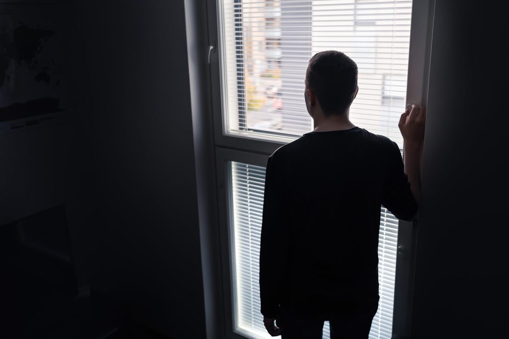 Image of a lonely man standing at a window looking out. Overcome your trauma symptoms and begin your healing journey with the help of EMDR therapy in Raleigh, NC.