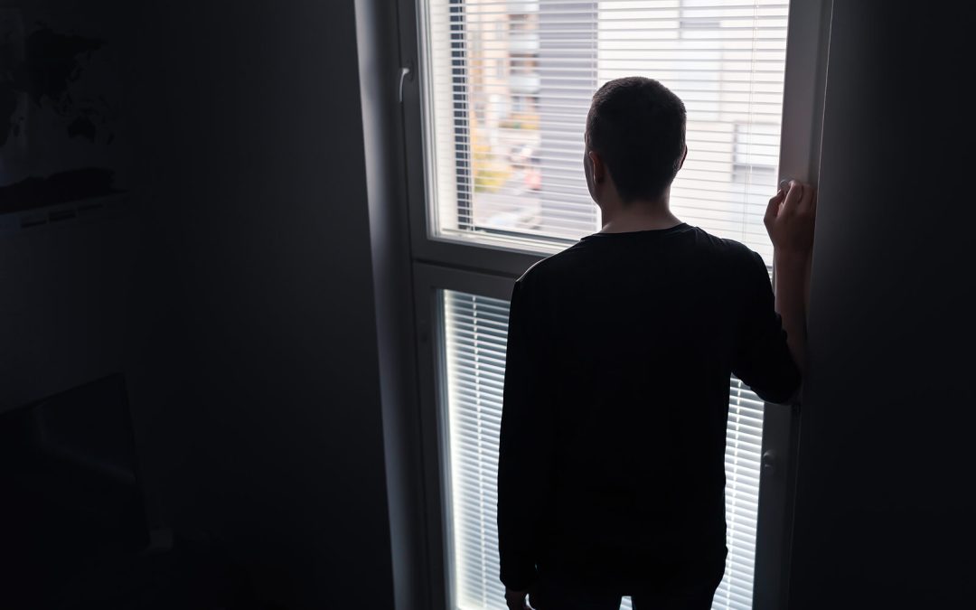 Image of a lonely man standing at a window looking out. Overcome your trauma symptoms and begin your healing journey with the help of EMDR therapy in Raleigh, NC.