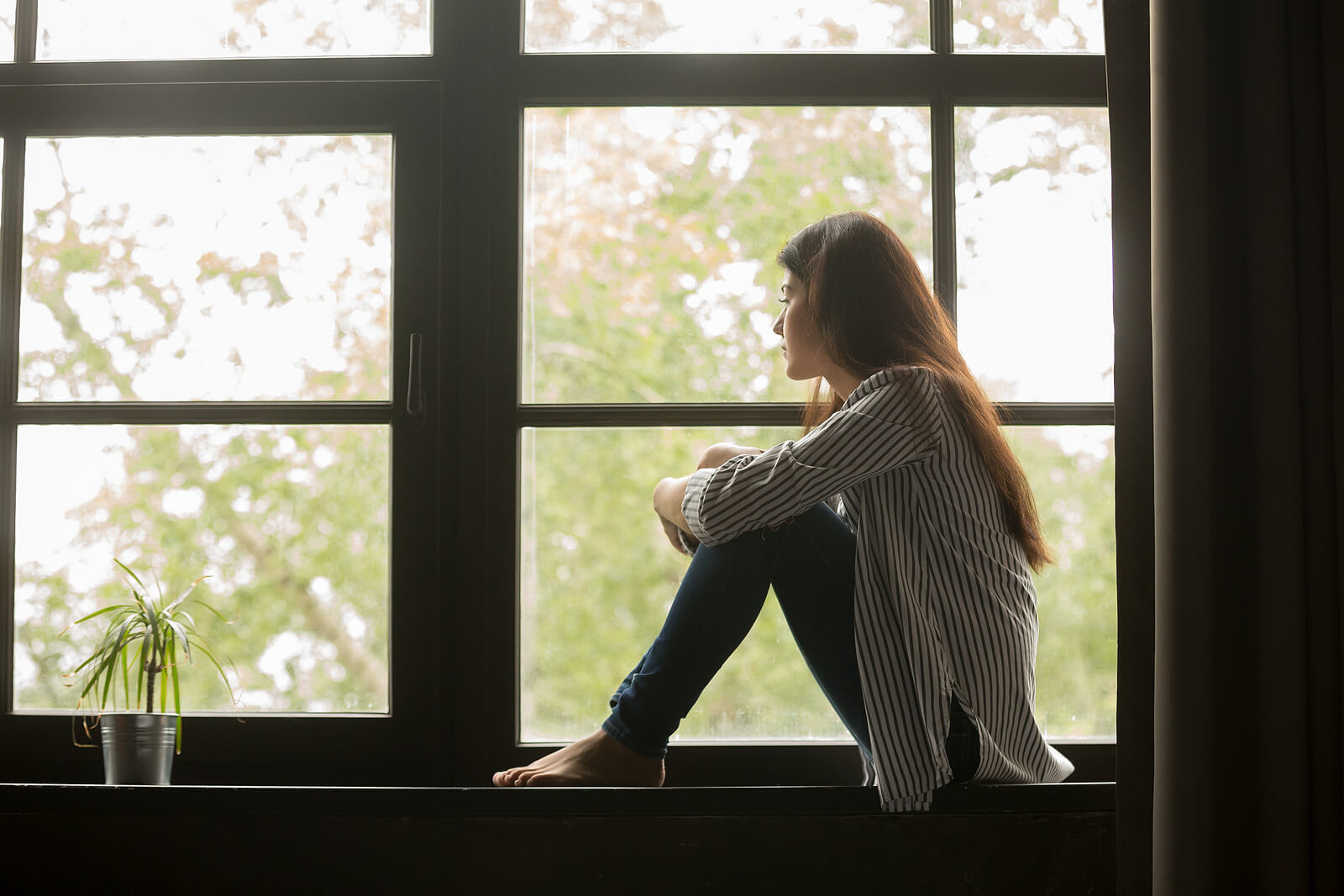 Image of a thoughtful woman sitting in a window sill looking out the window. If you find yourself struggling to heal from unresolved trauma, discover how EMDR therapy in Raleigh, NC can help you cope and start healing.