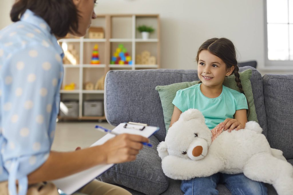 Image of a smiling little girl sitting on a couch speaking to a child EMDR therapist. Find support in helping your child heal and cope with their symptoms with the help of EMDR therapy in Raleigh, NC.