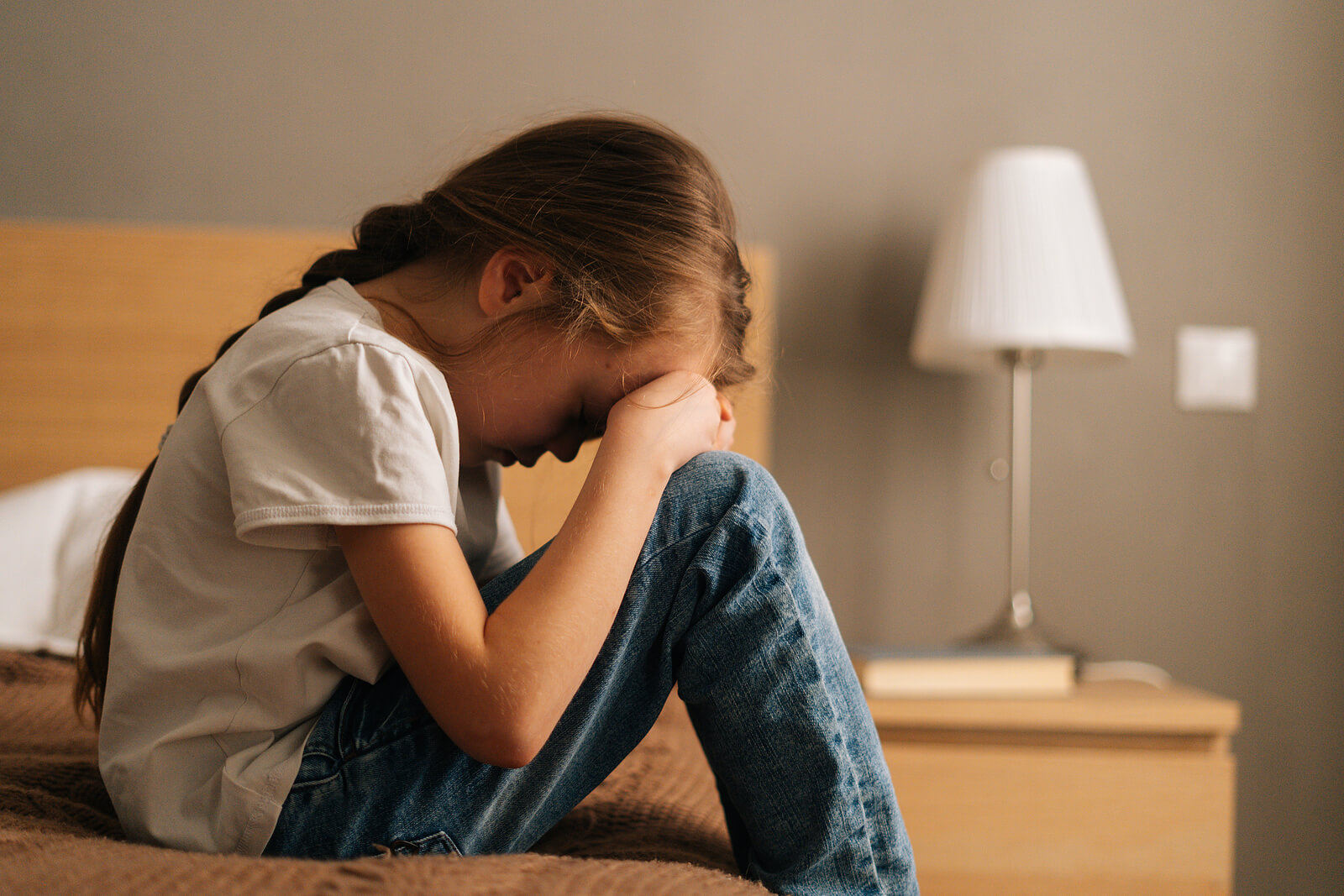 Anxious little girl sitting on a bed with her head in her hands. With the support of EMDR therapy for children in Raleigh, NC, your child can begin coping with their anxiety in healthy ways! Find support soon!