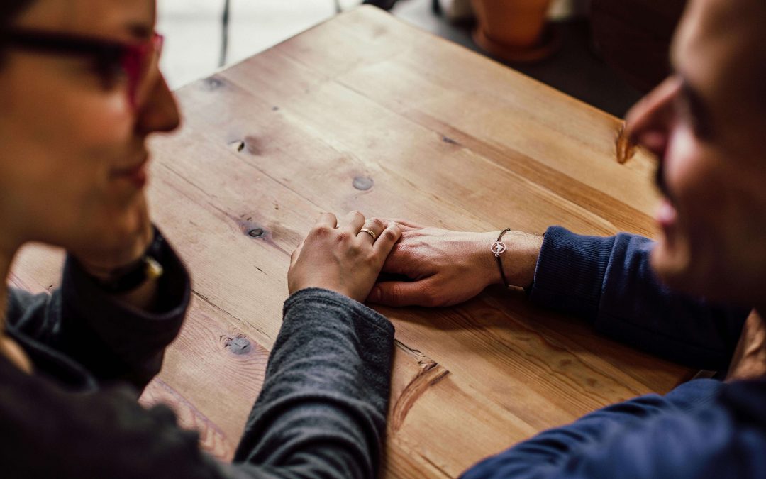 Image of a smiling couple sitting close at a table talking and holding hands. Learn to manage and cope with your emotional triggers due to trauma with the support of EMDR therapy in Raleigh, NC.